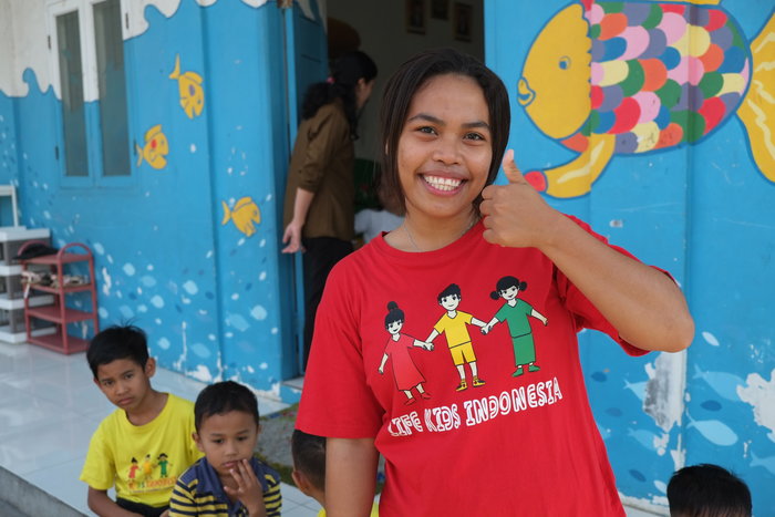 Ince smiles and puts her thumb up. In the background are the kindergarten school and some children.